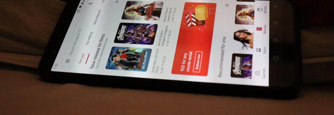 A smartphone displaying a streaming app interface, showing various movie and TV show thumbnails. The screen includes categories like 'For you' and 'New release hit films', with images from popular films. The background is a dimly lit setting with a fabric surface.