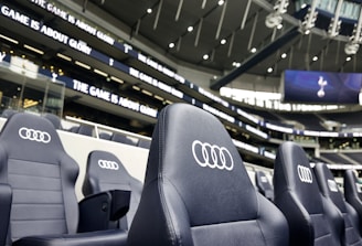 A row of black leather stadium seats with the Audi logo prominently displayed in an upscale sports venue. In the background, there are large screens and bright signage displaying the words 'The game is about glory,' suggesting a high-profile sporting event location.