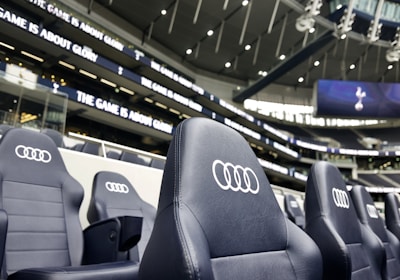A row of black leather stadium seats with the Audi logo prominently displayed in an upscale sports venue. In the background, there are large screens and bright signage displaying the words 'The game is about glory,' suggesting a high-profile sporting event location.