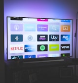 A flat-screen television is displayed in a dimly lit room, showcasing a variety of streaming service apps on its screen, such as Netflix, Prime Video, and Spotify. The TV is positioned on a black stand beside a tall floor lamp, casting a soft ambient light on the surrounding area.