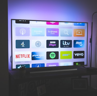 A flat-screen television is displayed in a dimly lit room, showcasing a variety of streaming service apps on its screen, such as Netflix, Prime Video, and Spotify. The TV is positioned on a black stand beside a tall floor lamp, casting a soft ambient light on the surrounding area.