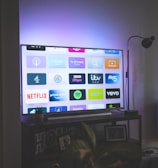 A flat-screen television is displaying a variety of streaming app icons, including Netflix, Prime Video, Spotify, and ITV Hub among others. The TV is placed on a media console with a small potted plant beside it. A tall lamp stands to the right, and a faint, ambient light casts a purple hue on the wall above the TV.