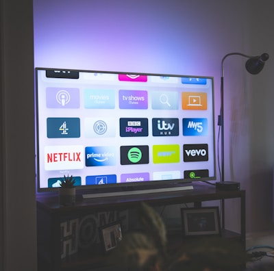 A flat-screen television is displaying a variety of streaming app icons, including Netflix, Prime Video, Spotify, and ITV Hub among others. The TV is placed on a media console with a small potted plant beside it. A tall lamp stands to the right, and a faint, ambient light casts a purple hue on the wall above the TV.