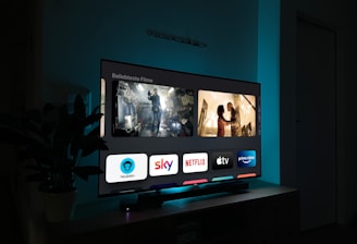 A television screen displaying a user interface with popular streaming service apps including Netflix, Sky, Apple TV, Prime Video, and others. The screen also shows images from movies or shows above the apps. The room is dimly lit with a potted plant visible on the side.