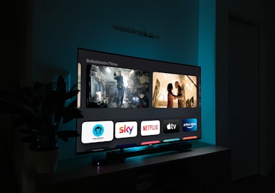 A television screen displaying a user interface with popular streaming service apps including Netflix, Sky, Apple TV, Prime Video, and others. The screen also shows images from movies or shows above the apps. The room is dimly lit with a potted plant visible on the side.