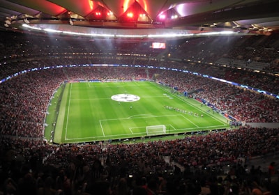 A large, packed stadium with bright lights illuminates a green soccer field. The scene is vibrant with a massive audience surrounding the pitch, visibly excited and engaged. The center of the field displays a prominent circular logo, suggesting a major sporting event. Players and staff can be seen on the field, likely preparing for a match.