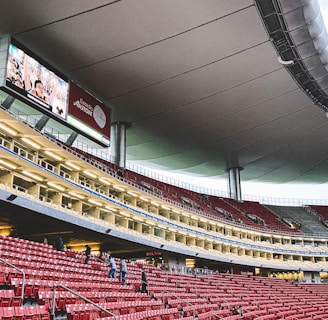 A large, modern stadium with red seats and a sleek, expansive roof structure. A few people are scattered among the seats, and there are large digital screens displaying advertisements. The setting is well-lit with multiple levels visible, suggesting a multi-tiered structure.