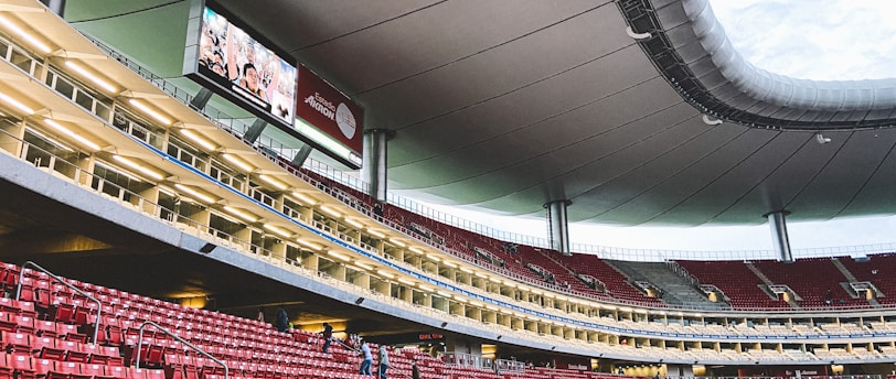 A large, modern stadium with red seats and a sleek, expansive roof structure. A few people are scattered among the seats, and there are large digital screens displaying advertisements. The setting is well-lit with multiple levels visible, suggesting a multi-tiered structure.