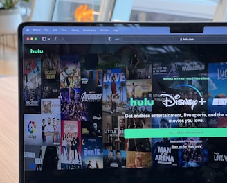 A laptop screen displays a web page featuring a mix of movie and TV show covers, highlighting popular titles and a Hulu and Disney+ bundle promotion. There are visible icons and images of films and shows alongside promotional text offering entertainment and live sports.