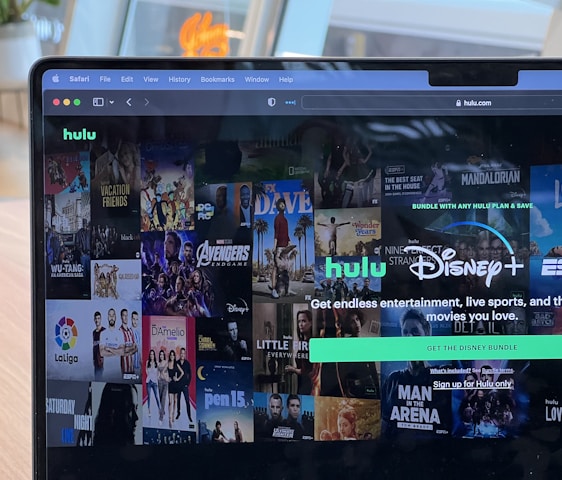 A laptop screen displays a web page featuring a mix of movie and TV show covers, highlighting popular titles and a Hulu and Disney+ bundle promotion. There are visible icons and images of films and shows alongside promotional text offering entertainment and live sports.