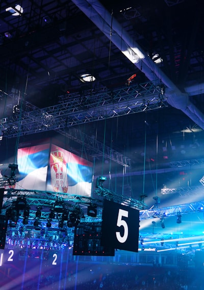 A large indoor arena is equipped with advanced lighting and audio systems. Suspended above the space are numerous digital displays showcasing numbers. The environment is filled with blue illumination and spotlights creating a vibrant atmosphere.