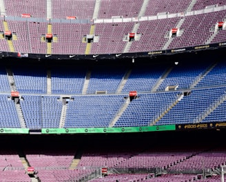 An empty football stadium with multiple tiers of seating. The seats are primarily purple in the upper sections and blue in the middle sections. There are several advertisement boards featuring brand names such as Spotify. Stairways and aisles are clearly marked, and the stadium appears well-maintained.