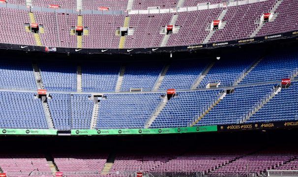 An empty football stadium with multiple tiers of seating. The seats are primarily purple in the upper sections and blue in the middle sections. There are several advertisement boards featuring brand names such as Spotify. Stairways and aisles are clearly marked, and the stadium appears well-maintained.