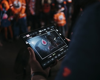 A person is holding a tablet that displays a video or image of a red and blue emblem on the screen. In the background, several people wearing dark and orange colored clothing, possibly sports jerseys, are partially visible. The scene is dimly lit, suggesting an indoor or evening setting.