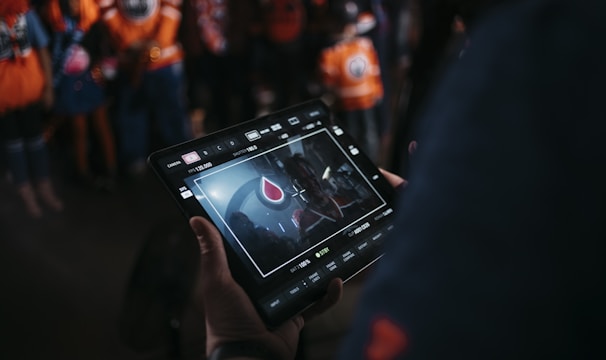 A person is holding a tablet that displays a video or image of a red and blue emblem on the screen. In the background, several people wearing dark and orange colored clothing, possibly sports jerseys, are partially visible. The scene is dimly lit, suggesting an indoor or evening setting.