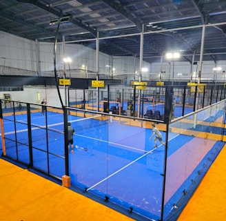 An indoor sports facility featuring several padel courts with blue playing surfaces and orange sidelines. There are players engaged in a game on one of the courts, while the others appear empty. The area is well-lit with overhead lighting and enclosed by a high ceiling and metal structure.