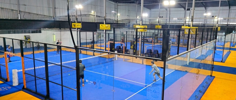 An indoor sports facility featuring several padel courts with blue playing surfaces and orange sidelines. There are players engaged in a game on one of the courts, while the others appear empty. The area is well-lit with overhead lighting and enclosed by a high ceiling and metal structure.