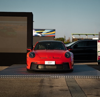 A bright red sports car is displayed on a raised platform outdoors, with a large digital screen on one side and another vehicle partially in view. The setting appears to be an automotive event or showcase, with clear blue skies in the background.