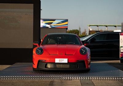 A bright red sports car is displayed on a raised platform outdoors, with a large digital screen on one side and another vehicle partially in view. The setting appears to be an automotive event or showcase, with clear blue skies in the background.