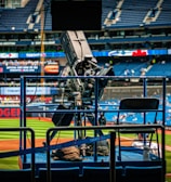 A professional-grade video camera is mounted on a platform overlooking a baseball field. The stadium seats in the background are filled with some spectators, and various advertisements are visible around the field. A staff member wearing a jacket with 'STAFF' written on the back is positioned near the camera.